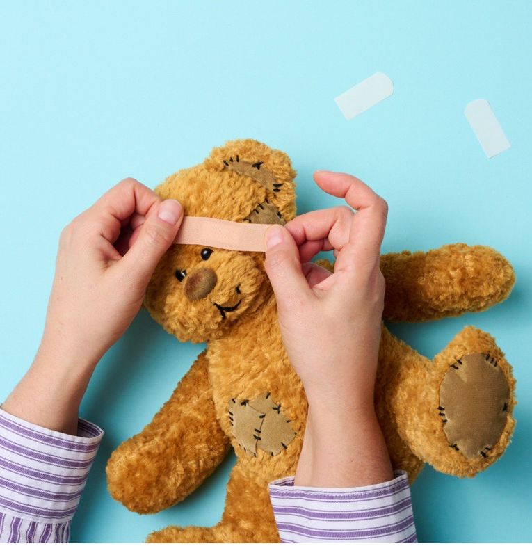 A pair of hands applying a bandage to a worn teddy bear, symbolising gentle healing and inner-child therapy.